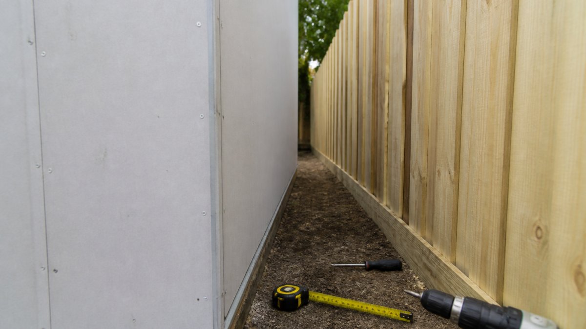 A close-up photograph of a newly built shed wall clad in fire-rated cement sheet, positioned close to a timber paling fence, with building tools and a tape measure resting against the base showing the narrow gap between wall and fence line